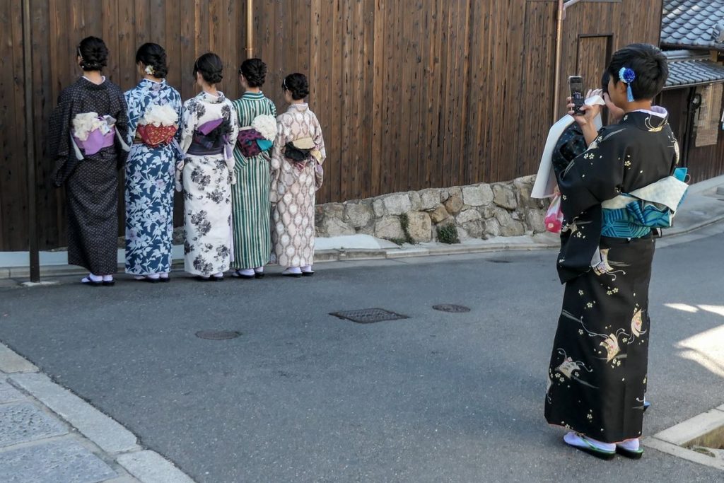 京都の通りで、着物姿で並ぶ友人たちを後ろからスマートフォンで撮影する人