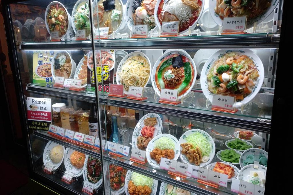 東京の飲食店の店頭で、ラーメンや炒め物、丼ものの食品サンプルが値札つきでガラスケースに並ぶ様子