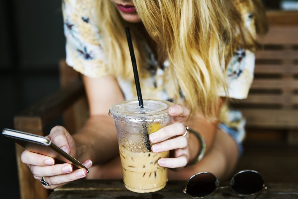 カフェのテーブルでアイスコーヒーを持ちながら、もう片方の手でスマートフォンを操作する手元