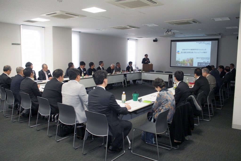 日本のセミナールームで、ロの字に並べた机に参加者が座り、前方のスクリーンを見ながら会議をしている様子