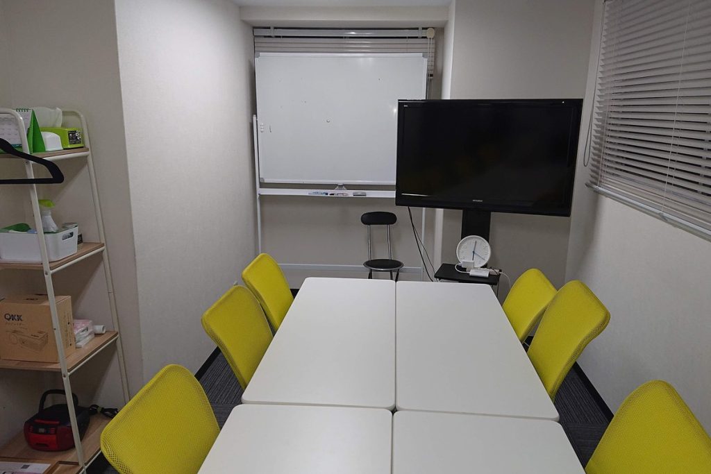 名古屋市中区の小さな会議室。白い机を黄色い椅子が囲み、奥にホワイトボードとモニターがある
