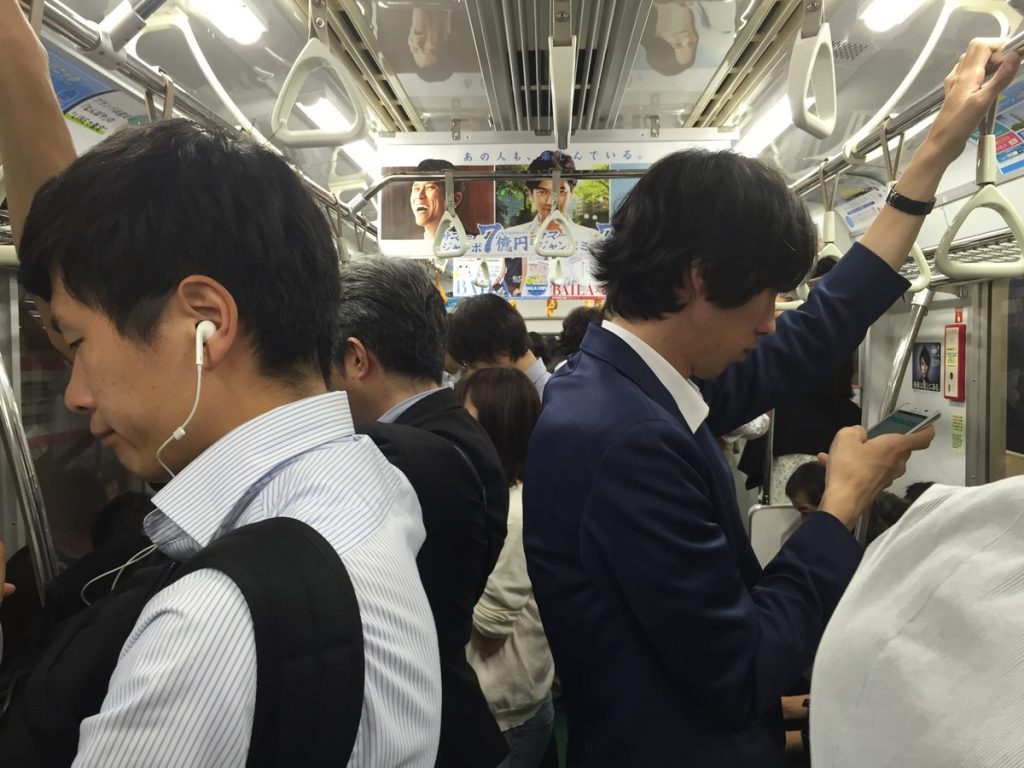 夜の混雑した東京の地下鉄車内で、立っている乗客がつり革やスマホを持っている様子